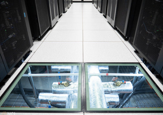 Clear floor tiles between two rows of tall black server racks reveal water pipes supplying cooling water for the Gadi supercomputer.