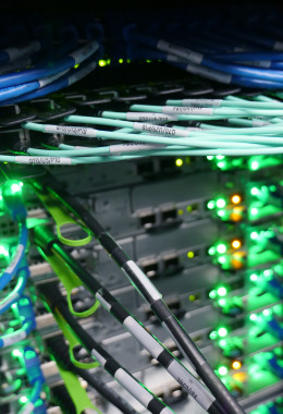 Close-up picture of a range of computer cables plugged into servers with bright green flashing lights, seen from above.