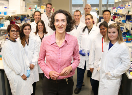 Sally Dunwoodie stands smiling at the camera surrounded by colleagues in white lab coats.