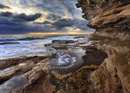 Striking clouds are a backdrop to swirling water in rock pools under layered rocky cliff walls.