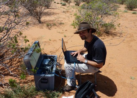 Graham Heinson sits on a stool in the desert in front of computer and other technical scientific equipment.