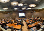Wide view of a full, beautiful auditorium with wooden desks and carpeted floor, with a woman speaking under a big projector screen saying "Welcome to ALCS2023".
