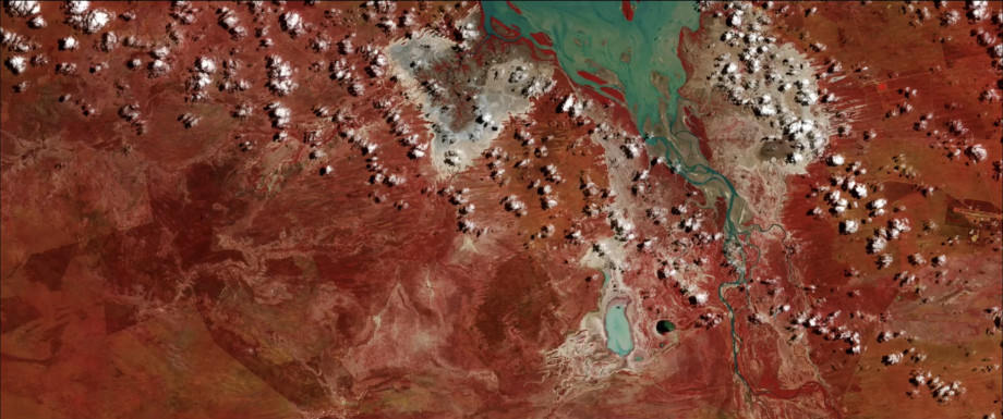 Screenshot from the Open Air video art project by Grayson Cooke. The video shows time lapse image sequences of the Australian landscape made using satellite imagery, interspersed with macro photography of paintings being created. The screenshot shows a river and lake in false colour blue, with lots of tiny clouds and the red outback.