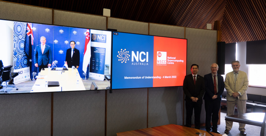 Two men visible standing behind a boardroom table on a large television, with another large television displaying the logos of NCI and NSCC on the right, and three men standing to the right of both televisions.