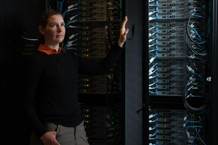 A woman stands in a dark space with her hand on the door of a supercomputer cabinet. She and the complex cables in the supercomputer are brightly lit.