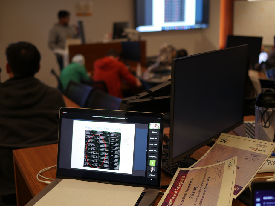 Screen of a laptop in focus with coding from a presentation visible, with other members of the audience and the presenter visible blurred further back in a dark seminar room.