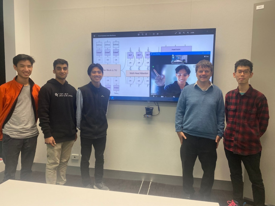 Group photo of men standing in front of a large television pointing to one remotely connected colleague on the screen. Everyone is posing for a photo.