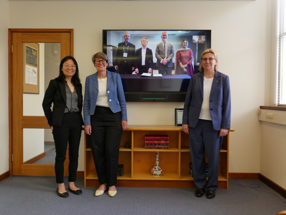 Group photo of three women standing in front of a large television showing their remotely connected colleagues on the screen. Everyone posing for a photo.