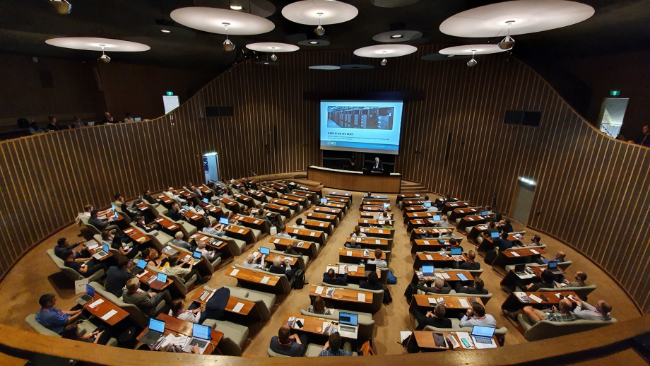 A view down into a circular auditorium with desks looking down towards a speaker in the middle of screen.