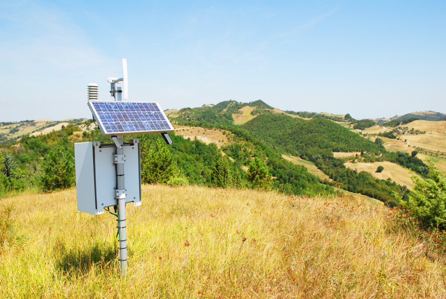 Weather station instruments on a grassy, sunny hillside. Hills and a blue sky are in the distance.