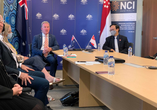 Two men sitting at the end of a long boardroom table, with Australian and Singaporean flags behind them and other people sitting further down the table.