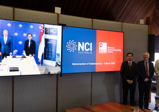 Two men visible standing behind a boardroom table on a large television, with another large television displaying the logos of NCI and NSCC on the right, and three men standing to the right of both televisions.