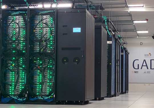 Long view of a data centre, with multiple pods of server racks all visible in a row. Bright green lights are visible on the nearest pod, and against the back wall is a sign that says Gadi in big letters.