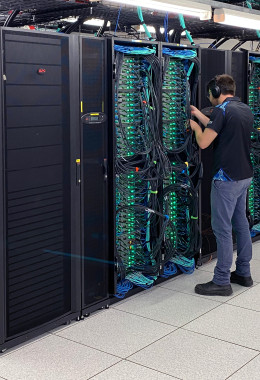 A technician working on the cabling in a pod of computer servers with green lights and complex cabling.