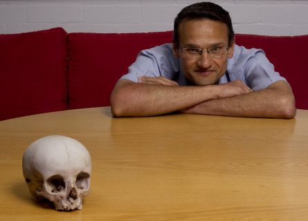 A human skull on a wooden table filling the frame, with a man behind the table leaning on it with arms crossed.