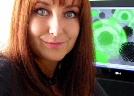 A woman, Amanda Barnard, smiles at the camera from in front of a computer showing nanomaterial structures.