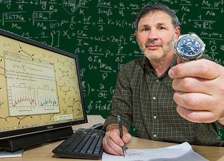 Anatoli Kheifets sits at a computer, in front of a blackboard filled with equations, holding a watch out to the camera.