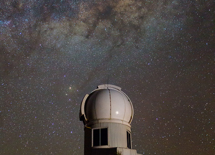 A round telescope dome sits on top of a hill under a brilliant night sky filled with thousands of stars in the Milky Way.