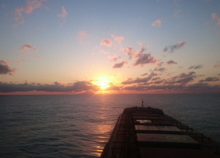 A container ship pointing towards the setting sun, with small bands of clouds near the horizon.