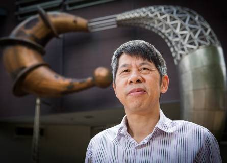 A man, Qinghua Qin, stands in front of an abstract sculpture showing two curved shapes reminiscent of biological creatures.