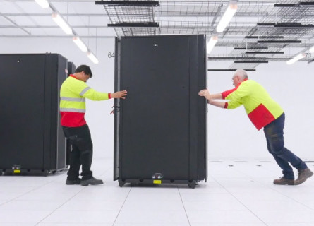 Two men roll a large black cabinet into position alongside other cabinets inside a large white room.