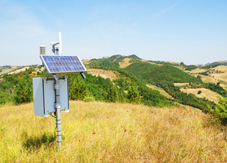 Weather station instruments on top of a grassy, sunny hillside, with forested hills in the distance. 