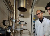 Two scientists stand looking at a set of scientific equipment with a bright orange flame burning in the centre.