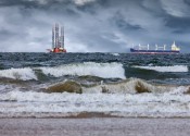 An oil rig and shipping container off shore from a beach while dark storm clouds approach.