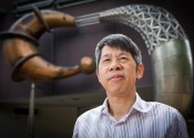 A man, Qinghua Qin, stands in front of an abstract sculpture showing two curved shapes reminiscent of biological creatures.