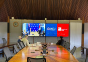 View of a long boardroom table, with three men seated at the end facing two televisions. The televisions are showing the other end of a video conference with people seated at a similar table visible on the screen on the left, and the logos of NCI and NSCC on the right.