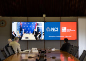Long view along a boardroom table. Three men are seated at the end of the table looking at television screens showing the NCI and NSCC logos, and the other attendees of the video conference they are participating in.