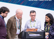 Four people standing at a lectern looking at a computer together. They are wearing lanyards and at a conference. The words OpenACC and Hackathons Asia-Pacific Summit 2022 are in a banner on the top of the image.