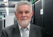 Close-up portrait of Sean Smith, a man with a short white beard wearing a suit and tie next to a high-tech looking server setup.