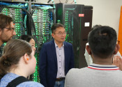 A man in a blue suit stands in front of a supercomputer with green lights. Facing him there are a number of people listening to him whose backs we can see.