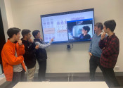 Group photo of men standing in front of a large television pointing to one remotely connected colleague on the screen. Everyone is posing for a photo.