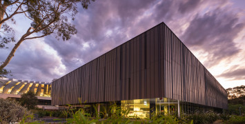 A rectangular building with striking vertical stripes in shades of grey and brown viewed from an angle, with hazy purple clouds above.