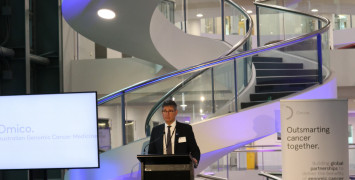 Professor David Thomas presenting on stage in front of an Omico banner in the Garvan Institute of Medical Research.