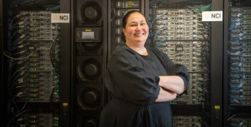 A woman in a black dress, Azure Hermes, stands with her arms crossed smiling at the camera. She is standing in front of a wall of computer servers with flashing lights and cables, with the NCI Australia logo visible.