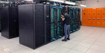 A technician working on the cabling in a pod of computer servers with green lights and complex cabling.
