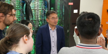 A man in a blue suit stands in front of a supercomputer with green lights. Facing him there are a number of people listening to him whose backs we can see.