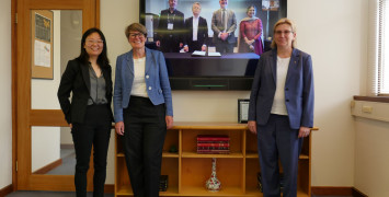 Group photo of three women standing in front of a large television showing their remotely connected colleagues on the screen. Everyone posing for a photo.